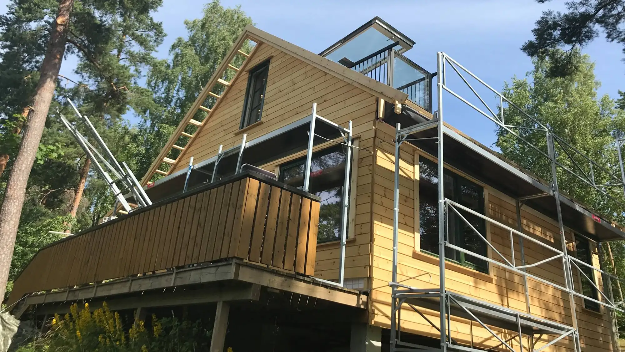 Trekledd bolig med balkong og stillas under oppføring i skogkant